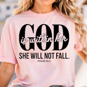 May include: A light pink t-shirt with the text "GOD is within her SHE WILL NOT FALL. Psalm 46:5" in black. The shirt has short sleeves and a relaxed fit.