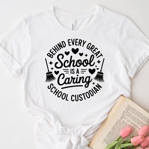 May include: White t-shirt with a black graphic that reads "Behind Every Great School Is a Caring School Custodian." The design includes hearts, stars, and brooms. A book and pink flowers are in the bottom right corner.