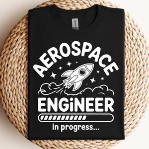 May include: Black t-shirt with a white graphic. The design includes the words "AEROSPACE ENGINEER", a rocket ship illustration, and the words "in progress..." below a progress bar.