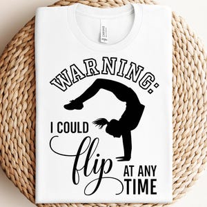Puede incluir: Camiseta blanca con texto negro que dice "Advertencia: Puedo hacer un flip en cualquier momento". El texto está en negrita, en una fuente divertida. Una silueta de una persona haciendo un salto mortal hacia atrás está sobre el texto.