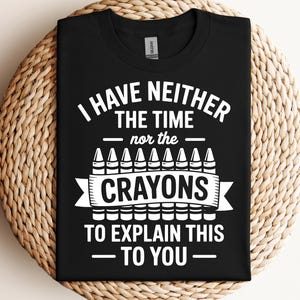 Puede incluir: Camiseta negra con el texto blanco "I HAVE NEITHER THE TIME nor the CRAYONS TO EXPLAIN THIS TO YOU". El diseño incluye ilustraciones de crayones. La camiseta se muestra sobre una superficie tejida.