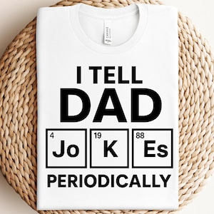 Peut inclure: T-shirt blanc avec l'inscription noire "I TELL DAD JOKES PERIODICALLY". Le mot "JOKES" est stylisé comme des éléments du tableau périodique. Le t-shirt est présenté sur une surface tissée.