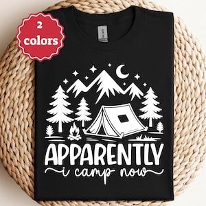 Peut inclure: T-shirt noir avec un motif de camping blanc. Le motif comprend une tente, des montagnes, des arbres, un feu de camp, des étoiles et un croissant de lune. Le texte dit "Apparently I camp now." Une étiquette rouge indique que le t-shirt est disponible en deux couleurs.