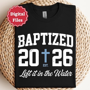 Może przedstawiać: Czarny t-shirt z białym, postarzanym napisem BAPTIZED 20 26, gdzie jasnoniebieski krzyż zastępuje zero. Napis EST. znajduje się pod krzyżem, a fraza Left it in the Water jest nadrukowana poniżej białą czcionką imitującą pismo odręczne.