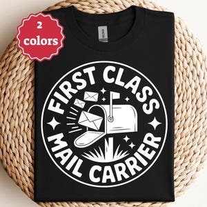 May include: Black t-shirt with a white graphic that reads "FIRST CLASS MAIL CARRIER" around a mailbox with envelopes. A red tag in the upper left corner says "2 colors". The shirt is displayed on a woven surface.