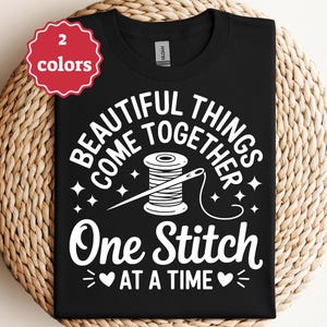Puede incluir: Camiseta negra con texto blanco: "BEAUTIFUL THINGS COME TOGETHER One Stitch AT A TIME". Presenta un carrete de hilo y una aguja. Una insignia roja y blanca de "2 colores" está en la esquina superior izquierda. La camiseta se muestra sobre una superficie tejida.