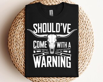 Puede incluir: Camiseta negra con un diseño de calavera y huesos cruzados blancos. El texto "Should've Come With A Warning" está impreso en la camiseta.