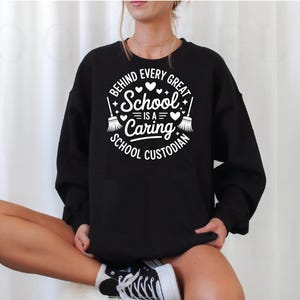 May include: Black sweatshirt with a white graphic that reads "BEHIND EVERY GREAT SCHOOL IS A CARING SCHOOL CUSTODIAN." The design includes hearts, stars, and brooms. The sweatshirt is long-sleeved and has a crew neck.