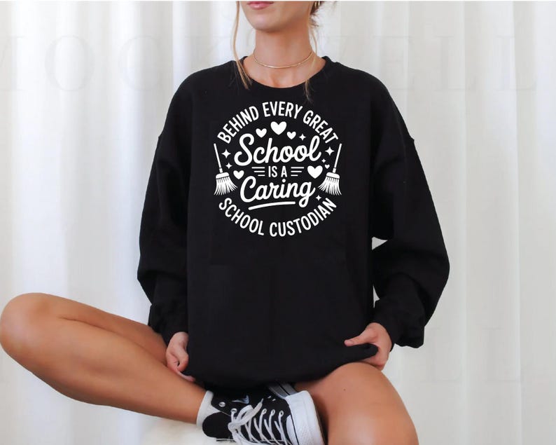 May include: Black sweatshirt with a white graphic that reads "BEHIND EVERY GREAT SCHOOL IS A CARING SCHOOL CUSTODIAN." The design includes hearts, stars, and brooms. The sweatshirt is long-sleeved and has a crew neck.