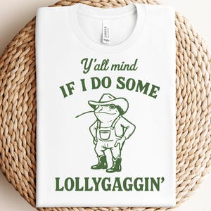 Puede incluir: Camiseta blanca con un diseño verde de una rana con sombrero de vaquero y peto. El texto dice "Y'all mind if I do some lollygaggin'." La camiseta está sobre una superficie tejida de color marrón claro.