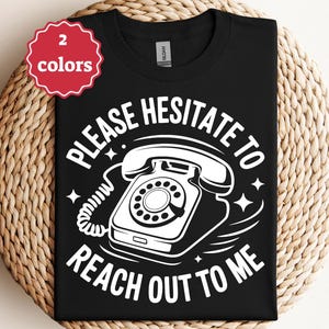 May include: Black t-shirt with a white graphic of a rotary phone and the text "PLEASE HESITATE TO REACH OUT TO ME". A red and white tag indicates the shirt is available in two colors.