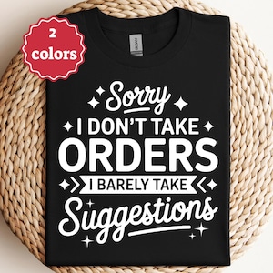 Op de afbeelding: Zwart T-shirt met witte tekst: "Sorry I don't take orders I barely take suggestions." Een rood-wit rond label in de linkerbovenhoek geeft twee beschikbare kleuren aan. Het T-shirt wordt gepresenteerd op een geweven oppervlak.