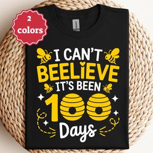 May include: Black t-shirt with a yellow and white graphic. The graphic reads "I can't beelieve it's been 100 days" with bee and honeycomb illustrations. A red and white tag in the upper left corner indicates "2 colors".