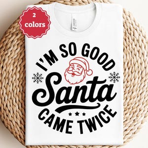 May include: White t-shirt with a black graphic that reads "I'M SO GOOD Santa CAME TWICE" with a Santa Claus face and snowflakes. A red and white sticker in the upper left corner says "2 colors". The shirt is displayed on a woven surface.