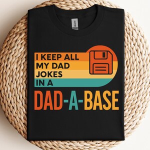 May include: Black t-shirt with the text "I KEEP ALL MY DAD JOKES IN A DAD-A-BASE" in orange, teal, and yellow. The design includes a retro-style floppy disk graphic. The shirt is displayed on a woven surface.