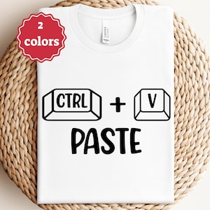 Könnte beinhalten: Weißes T-Shirt mit einer schwarzen Grafik der CTRL- und V-Tasten und dem Wort PASTE darunter. Ein rot-weißes Abzeichen in der oberen linken Ecke zeigt zwei verfügbare Farben an. Das T-Shirt wird auf einer gewebten Oberfläche präsentiert.