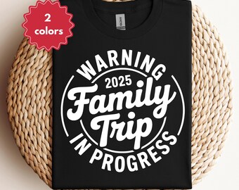 Può includere: T-shirt nero con grafica circolare bianca con la scritta "WARNING 2025 Family Trip IN PROGRESS". Un'etichetta rossa nell'angolo in alto a sinistra indica "2 colori". La maglietta è esposta su una superficie tessuta.