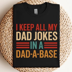 May include: Black t-shirt with the text "I KEEP ALL MY DAD JOKES IN A DAD-A-BASE" in red, beige, teal, and orange. The shirt is displayed on a woven, circular surface.