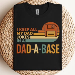 Puede incluir: Camiseta negra con el texto "I KEEP ALL MY DAD JOKES IN A DAD-A-BASE" en naranja, amarillo y verde azulado. El diseño incluye un gráfico de disquete de estilo retro. La camiseta se muestra sobre una superficie tejida.