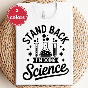 May include: White t-shirt with the text "STAND BACK I'M DOING Science" in black. The design includes laboratory glassware and stars. A red tag in the upper left corner indicates "2 colors". The shirt is displayed on a woven surface.