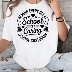 May include: White t-shirt with a black graphic that reads "BEHIND EVERY GREAT SCHOOL IS A CARING SCHOOL CUSTODIAN." The design includes hearts, stars, and broom illustrations. The shirt is a crew neck and has short sleeves.