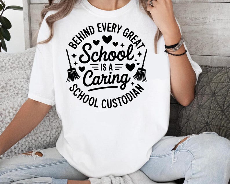 May include: White t-shirt with a black graphic that reads "BEHIND EVERY GREAT SCHOOL IS A CARING SCHOOL CUSTODIAN." The design includes hearts, stars, and broom illustrations. The shirt is a crew neck and has short sleeves.