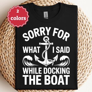 May include: Black t-shirt with a white anchor design and the text "SORRY FOR WHAT I SAID WHILE DOCKING THE BOAT." A red and white tag indicates the shirt is available in two colors. The shirt is displayed on a woven surface.