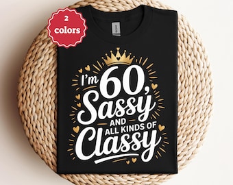 以下が含まれることがあります： 「I'm 60, Sassy and All Kinds of Classy」の文字が白と金色の黒いTシャツ。金の王冠とハートのアクセントも特徴です。赤いタグは、シャツが2色で利用可能であることを示しています。