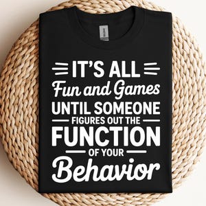 May include: Black t-shirt with the white text "IT'S ALL Fun and Games UNTIL SOMEONE FIGURES OUT THE FUNCTION OF YOUR Behavior". The shirt is displayed on a woven, circular surface.