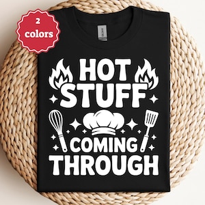 Puede incluir: Camiseta negra con el texto blanco "HOT STUFF COMING THROUGH", acompañada de ilustraciones de llamas, un gorro de chef, un batidor y una espátula. Una pegatina roja indica "2 colores".