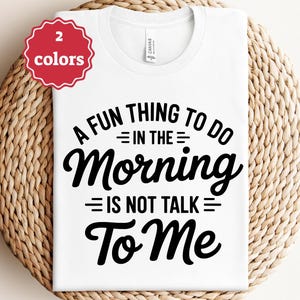 Puede incluir: Camiseta blanca con el texto negro "A FUN THING TO DO IN THE MORNING IS NOT TALK TO ME". Una insignia roja en la esquina superior izquierda indica "2 colores". La camiseta se muestra sobre una superficie tejida.