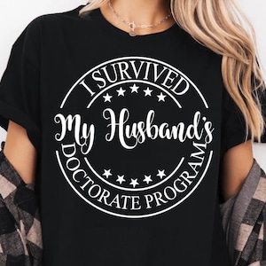 May include: Black t-shirt with a white circle graphic that says "I Survived My Husband's Doctorate Program".