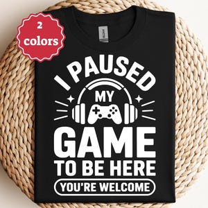 Puede incluir: Camiseta negra con un gráfico blanco que dice "I PAUSED MY GAME TO BE HERE YOU'RE WELCOME". El diseño incluye auriculares y un mando de juego. Una etiqueta roja y blanca en la esquina superior izquierda dice "2 colores".
