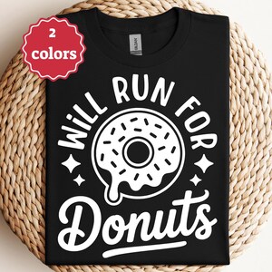 Könnte beinhalten: Schwarzes T-Shirt mit dem weißen Text "WILL RUN FOR DONUTS" und einer Donut-Grafik. Das Shirt wird auf einer gewebten Oberfläche präsentiert, mit einem rot-weißen Etikett, das zwei Farboptionen anzeigt.