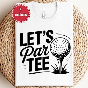 Pode incluir: Camiseta branca com o texto preto "LET'S Par TEE" e um gráfico de bola de golfe. Um adesivo vermelho no canto superior esquerdo indica "2 cores". A camiseta é exibida em uma superfície tecida.