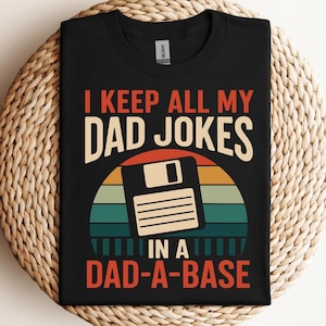 Op de afbeelding: Zwart T-shirt met de tekst "I KEEP ALL MY DAD JOKES IN A DAD-A-BASE" in wit, oranje en teal. Het ontwerp bevat een retro-stijl afbeelding van een diskette over een zonsondergang ontwerp. Een leuke, novelty tee.
