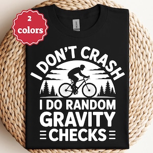 以下が含まれることがあります： 白文字とサイクリストのグラフィックが入った黒いTシャツ。「I DON'T CRASH I DO RANDOM GRAVITY CHECKS」と書かれています。赤と白のタグは、このシャツが2色で利用可能であることを示しています。シャツは織られた表面に表示されています。