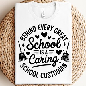 May include: White t-shirt with a crew neck featuring a black graphic. The graphic reads "BEHIND EVERY GREAT SCHOOL IS A CARING SCHOOL CUSTODIAN" with hearts, stars, and brooms. The shirt is displayed on a woven surface.