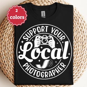 Op de afbeelding: Zwart T-shirt met een witte afbeelding met de tekst "Support Your Local Photographer". Het ontwerp bevat een camera, handen en een filmrol. Een rood-wit label geeft aan dat het shirt in twee kleuren verkrijgbaar is. Het shirt wordt gepresenteerd op een geweven oppervlak.