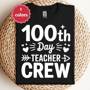 Może przedstawiać: Czarny t-shirt z białym napisem "100th Day Teacher Crew". Projekt zawiera serca, strzałki i gwiazdy. Czerwono-biała etykieta w lewym górnym rogu mówi "2 kolory".