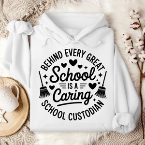 May include: A white hooded sweatshirt with the text "BEHIND EVERY GREAT SCHOOL IS A CARING SCHOOL CUSTODIAN" in a black circular design. The design includes hearts, stars, and broom illustrations. A white mug and snowflake cookie are on a wooden tray.