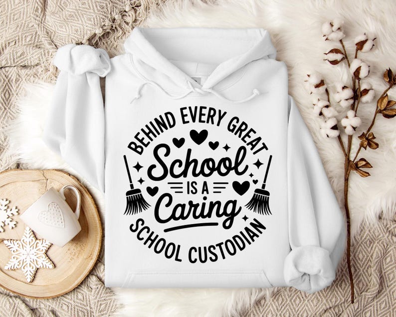 May include: A white hooded sweatshirt with the text "BEHIND EVERY GREAT SCHOOL IS A CARING SCHOOL CUSTODIAN" in a black circular design. The design includes hearts, stars, and broom illustrations. A white mug and snowflake cookie are on a wooden tray.