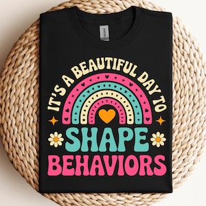 May include: Black t-shirt with a colorful rainbow design and the text "It's a beautiful day to shape behaviors." The design includes a heart, stars, and flowers. A fun and educational tee.
