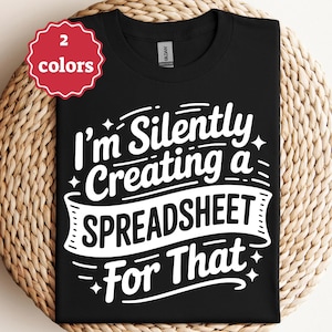Könnte beinhalten: Schwarzes T-Shirt mit dem weißen Text "I'm Silently Creating a Spreadsheet For That." Ein rot-weißes Etikett in der oberen linken Ecke zeigt "2 Farben". Das T-Shirt wird auf einer gewebten, kreisförmigen Oberfläche präsentiert.