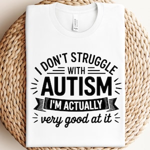 Op de afbeelding: Wit t-shirt met de tekst "I don't struggle with autism, I'm actually very good at it" in het zwart. Het shirt wordt gepresenteerd op een geweven, lichtbruin oppervlak.