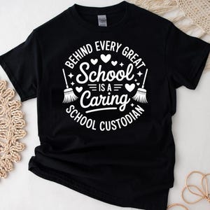 May include: Black t-shirt with a white graphic that reads "BEHIND EVERY GREAT SCHOOL IS A CARING SCHOOL CUSTODIAN." The design includes hearts and brooms. The shirt is made of a soft material.