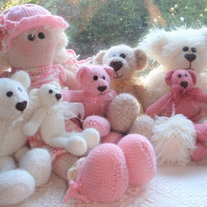May include: A group of knitted teddy bears in various shades of pink and white. The bears are arranged in a row on a white surface. Some of the bears have pink noses and eyes, while others have black noses and eyes. The bears are all wearing different outfits, including a pink dress, a white dress, and a pink and white striped shirt.
