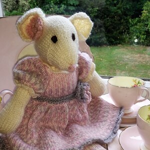 May include: A knitted mouse doll in a pink and grey dress and shoes. The doll has cream-coloured ears and a pink nose. Two teacups and saucers are in the background, with a garden visible through a window.