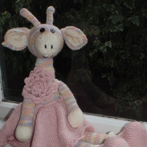 May include: A knitted giraffe doll wearing a pink dress with a flower detail. The giraffe has a striped neck and pink and white legs.