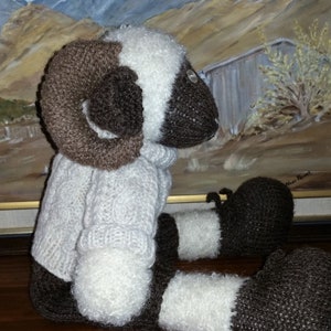 Op de afbeelding: Een gebreid pluche speelgoed rammetje met bruine hoorns, witte vacht en bruine poten. Het rammetje zit op een houten oppervlak.